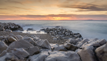 The sun sets over the beach in Petrčane, Croatia, with steely colored rocks in the foreground and the waves of the Adriatic Sea misty in the evening light with a long exposure effect.