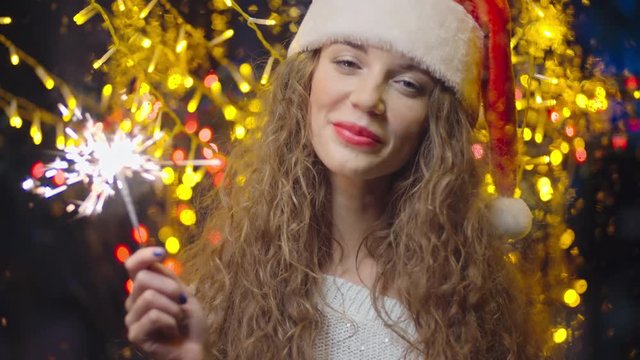 Young beautiful woman with long curly hair and Santa hat holding sparker and looking at camera with smile