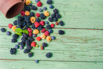 Multi-colored summer berries, poured out of the ceramic mug, on a rustic wooden table.