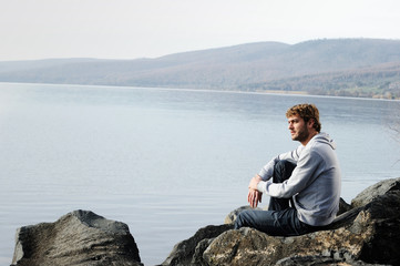 Fototapeta na wymiar Giovane uomo seduto su una roccia in riva al lago