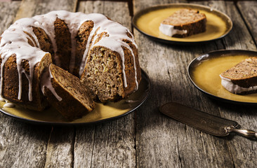 Banana cake with apples and walnuts drizzled frosting and sliced pieces on plates with effect craquelure on the shabby wooden background. Selective focus 