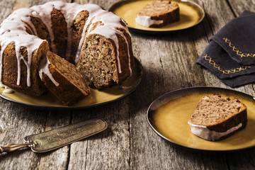Banana cake with apples and walnuts drizzled frosting and sliced pieces on plates with effect craquelure on the shabby wooden background. Selective focus 