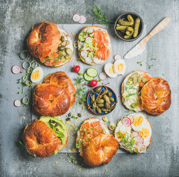 Variety of bagels with smoked salmon, eggs, radish, avocado, cucumber, greens and cream cheese for breakfast, healthy lunch or party over grey concrete background, top view. Takeaway food concept