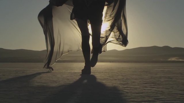 Girl with a fabric runs towards sunset in the desert. Young woman running towards sunrise, slow motion