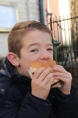 A handsome young boy eating a beef burger while outside during the daytime, 2016