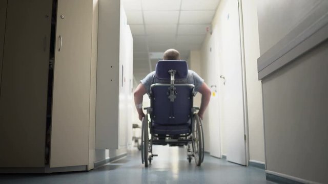 Rear view of disabled man pushes himself in wheelchair down hospital corridor