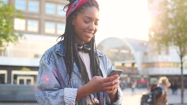 Black young woman typing on smart phone in Berlin. Smiling girl wearing a jeans jacket with lurred people and tram in Alexanderplatz on background.
