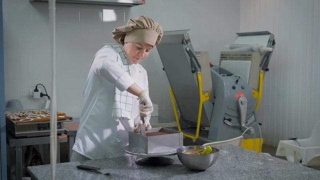 Serious woman standing behind povarsikim pastry table on the background of mechanical harvester confectionery and chocolate cake shovel cuts from metal mold and then removing it