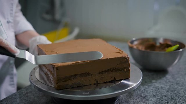 Kitchen in a candy store. The woman in kitchen cooks chocolate cake. The confectioner in gloves and a white form has prepared layers for cake and covers cake layers by means of the pallet