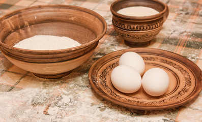 Fototapeta na wymiar Milk, three eggs, flour in a ceramic clay dish with traditional Ukrainian patterns and ornaments on a table with flour with a green tablecloth in a cage. The process of preparation of the dough