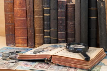 The old coins, banknotes, magnifying glass and open antiquarian book. - obrazy, fototapety, plakaty