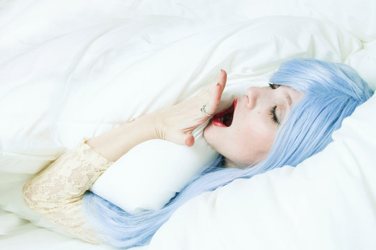 Chica joven con el pelo azul metida en su cama bostezando 