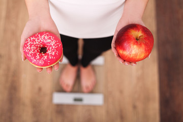 Diet. Woman Measuring Body Weight On Weighing Scale Holding Donut and apple. Sweets Are Unhealthy...