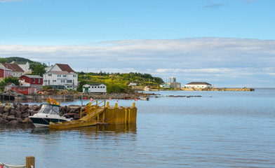 Fototapeta premium Golden adirondack chairs on a rock jetty. Houses on the sea along a village shoreline. Rural Newfoundland, Canada. Docked speedboat waits in silence.