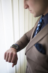 Close up of young businessman in a brown suit standing in his office while looking at his smart watch.