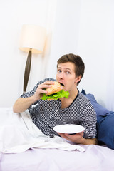 Shocked single man eating hamburger while lying in bed. Handsome man looking at camera and having breakfast in morning at home.