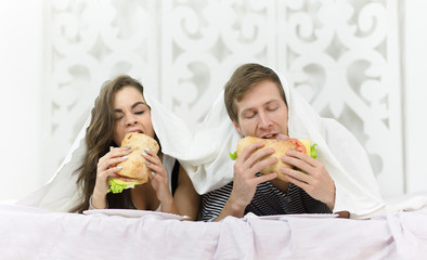 Young couple eating delicious hamburgers while lying in bed covered. People resting and relaxing...