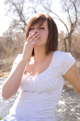 Cute brunette smokes a cigarette outside next to a river. Puts the cigarette next to her mouth and enhales.