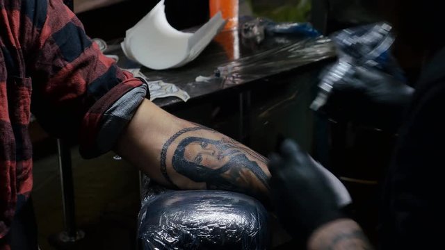 Master tattoo draws on the clients tattoo. Professional artist holding a black tattoo machine in black sterile gloves and working, makes a tattoo on a male hand