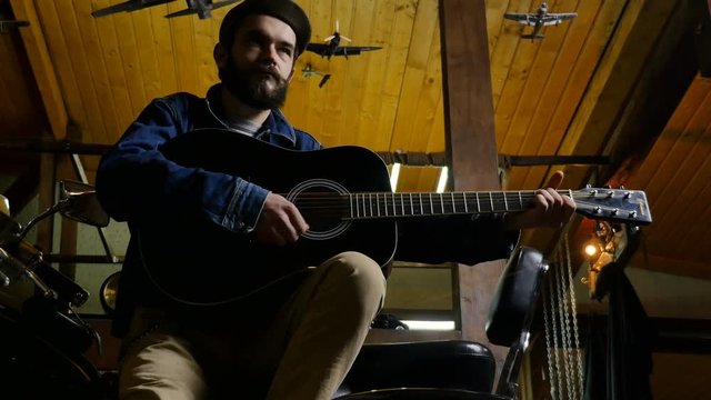 Biker plays guitar in the room with the aircraft on the ceiling. young bearded musician sitting on a motorcycle and playing guitar