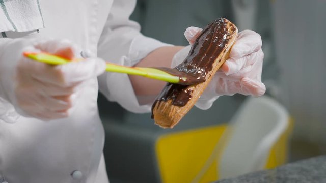 Confectioner gently smears thick layer chocolate on freshly baked eclairs. Professional chef during her favorite work. Drop of liquid cream fell down.