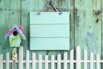 Blank mint green sign hanging by birdhouse and white picket fence