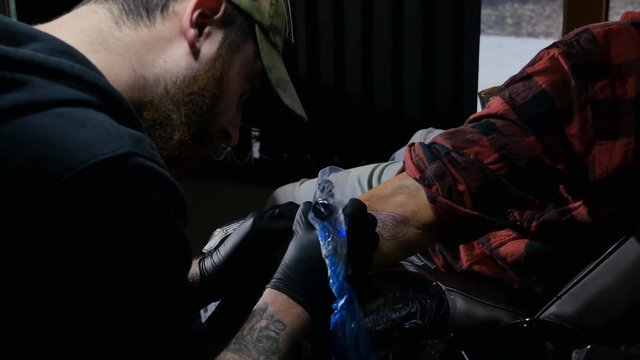 Bearded tattoo artist demonstrates close up the process of getting black tattoo with paint. Master works in black sterile gloves in art salon