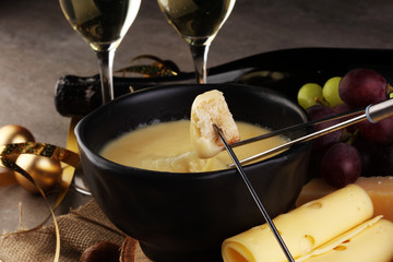 Gourmet Swiss fondue dinner on a winter evening with assorted cheeses on a board alongside a heated pot of cheese fondue with two forks dipping bread and white wine behind in a tavern or restaurant
