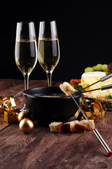 Gourmet Swiss fondue dinner on a winter evening with assorted cheeses on a board alongside a heated pot of cheese fondue with two forks dipping bread and white wine behind in a tavern or restaurant