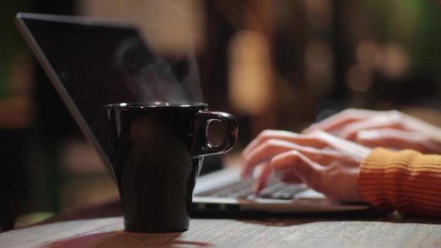 Footage of a person typing on a computer near a steamy coffee mug.