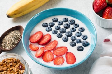 Yogurt fruit smoothie bowl with strawberries and blueberries. Jar of homemade granola, chia seeds, fresh banana and more strawberries on side. Bright morning shot