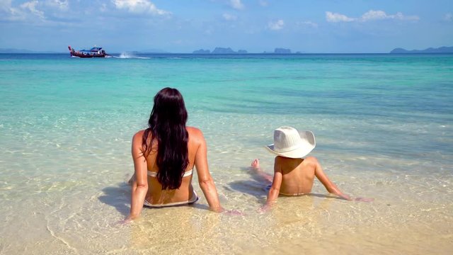 A mother and her son relaxing on a beach on the island Koh Kradan in southern Thailand watching a longtail boat pass by.