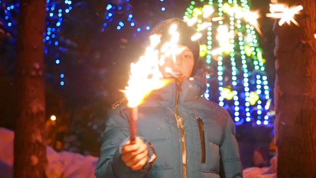 The child holds the sparklers outdoors in the winter. Slowmotion . In the background, lights and garlands of Christmas fir
