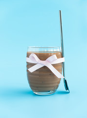 Homemade Banana, Almond Milk and Espresso Smoothie with Medjool Dates in a glass with pink bow and a drinking spoon on the side on pale blue background.