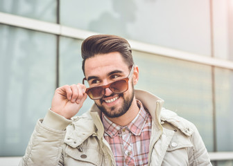 young guy with a beard and mustache with glasses in a suit posing on the street in the sunlight