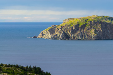 Naklejka premium Late afternoon sun shines on an outcropping of high rugged terrain in calm sea water in the bay at Twillingate, Newfoundland, Canada.