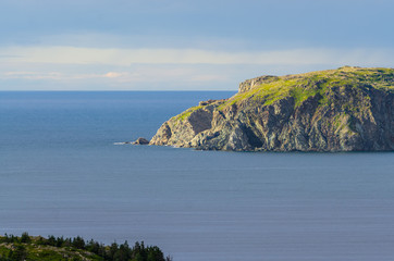 Late afternoon sun shines on an outcropping of high rugged terrain in calm sea water in the bay at Twillingate, Newfoundland, Canada.