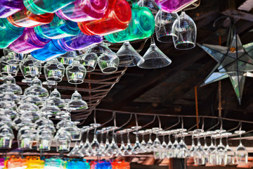 Restaurant interior background with narrow focus. Empty transparent wineglasses, rows of colorful beverage glasses hanging on top rack above bar counter. Vintage style wood ceiling.