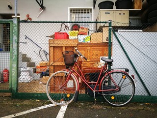 Rotes Damenrad mit schwarzem Gepäckkorb vor einem Hinterhof mit Drahtzaun, alten Möbeln und...
