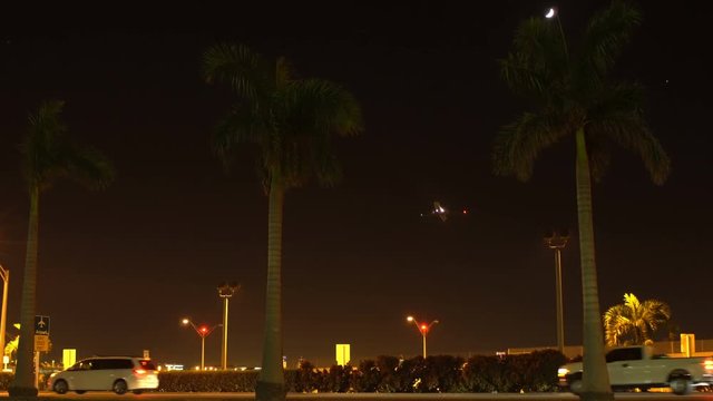 Wide angle night scene shot of a jumbo jet airplane taking off from behind the palm trees in the distance and flying across the dark sky above the highway