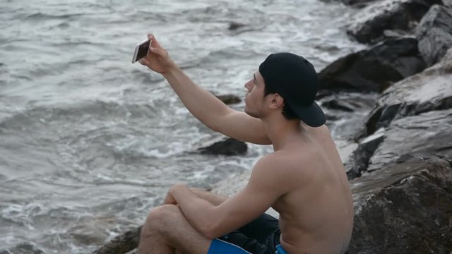 Shirtless Young Handsome Man Busy with his Mobile Phone Taking Selfie Photo While Sitting on Beach Boulders next to Sea or Ocean