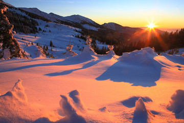 The setting sun lights snow-covered slopes of the mountains. Red lit areas and blue shadows.