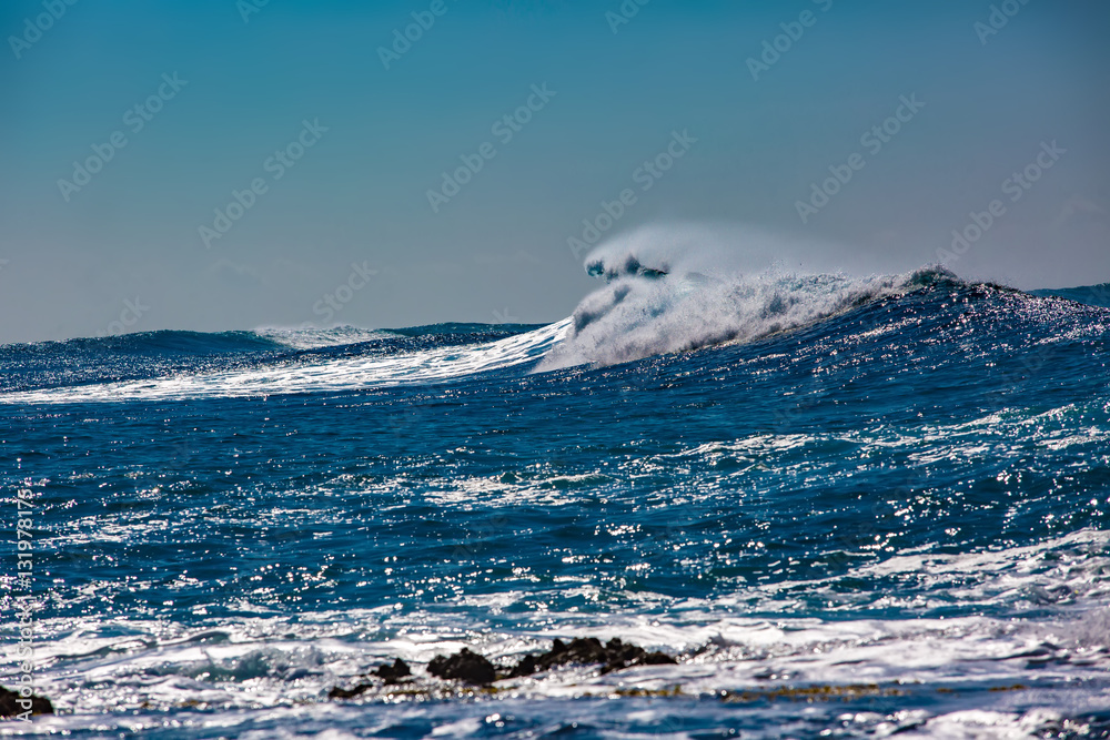 Wall mural Rough blue ocean wave crashing. Black rocks showing up from white foam.