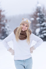 Attractive smiling young blonde girl walking in winter forest. Pretty woman in wintertime outdoor. Wearing winter clothes. Knitted sweater, scarf, hat and mittens.