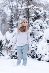 Fototapeta na wymiar Attractive smiling young blonde girl walking in winter forest. Pretty woman in wintertime outdoor. Wearing winter clothes. Knitted sweater, scarf, hat and mittens.
