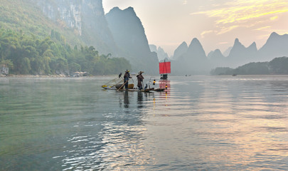 XINGPING, CHINA - OCTOBER 22, 2014: Fisherman stands on traditional bamboo boats at sunrise (boat with a red sail in the background) - The Li River, Xingping, China - 131983971