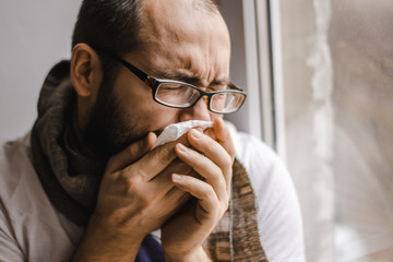 Young man blowing his nose while sitting at the window