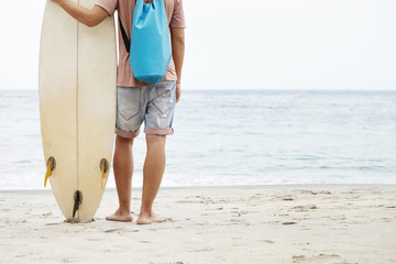 Healthy lifestyle and leisure concept. Rear cropped view of Caucasian tourist standing barefooted on sandy beach and holding white surfboard, facing calm and peaceful ocean, waiting for waves