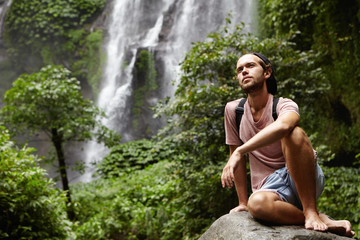 People, nature and adventure. Young hipster with backpack sitting on big rock by waterfall on background, having joyful look. Stylish barefooted hiker relaxing on stone during journey in jungle