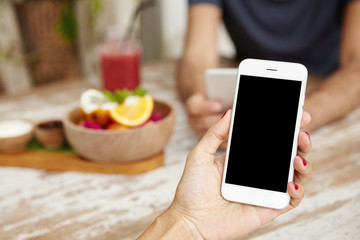 Cropped shot of female hand with red nails reading message via email or making call on generic smart phone, having lunch with her husband, sitting at wooden table in cozy interior. Film effect